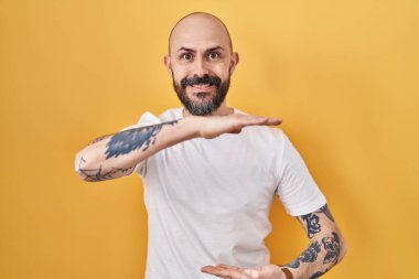 Young hispanic man with tattoos standing over yellow background gesturing with hands showing big and large size sign, measure symbol. smiling looking at the camera. measuring concept. 
