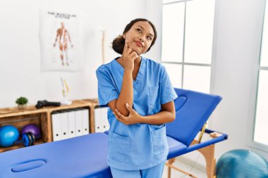Güzel İspanyol fizyoterapist kadın ağrı kesici kliniğinde elleri çenesinde soru sorarken, düşünceli bir ifade takınıyor. Düşünceli bir yüzle gülümsüyor. şüphe kavramı. 