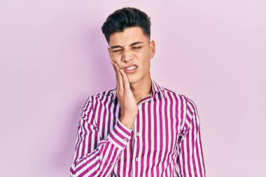 Günlük kıyafetler giyen genç İspanyol adam diş ağrısı ya da diş hastalığından dolayı acı dolu bir ifadeyle ağıza dokunuyor. dişçi 