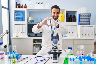 Sakallı genç bir İspanyol, bilim adamının laboratuvarında çalışıyor. Sevgiyle gülümsüyor, elleriyle kalp sembolü yapıyor. romantik kavram. 