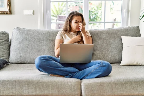 Beautiful young brunette woman sitting on the sofa using computer laptop at home smelling something stinky and disgusting, intolerable smell, holding breath with fingers on nose. bad smell