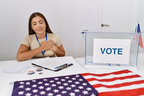 Young brunette woman at political election sitting by ballot smiling with hands on chest with closed eyes and grateful gesture on face. health concept.