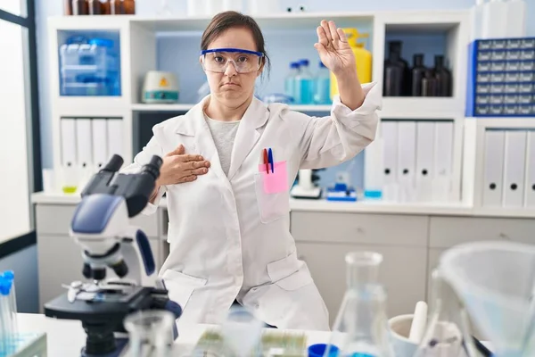 Hispanic girl with down syndrome working at scientist laboratory swearing with hand on chest and open palm, making a loyalty promise oath