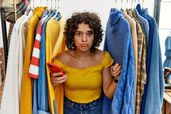 Young hispanic woman searching clothes on clothing rack using smartphone depressed and worry for distress, crying angry and afraid. sad expression.