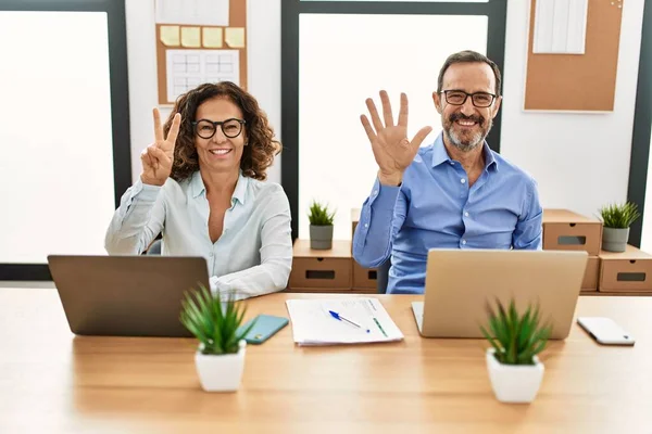 Middle age hispanic woman and man sitting with laptop at the office showing and pointing up with fingers number seven while smiling confident and happy.