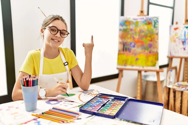 Young brunette teenager at art studio with a big smile on face, pointing with hand finger to the side looking at the camera.