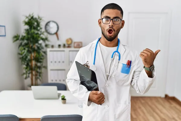 Young Indian Man Wearing Doctor Uniform Stethoscope Surprised Pointing Hand — Stockfoto