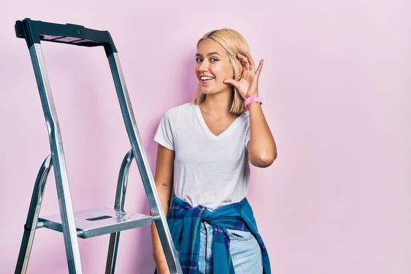 Beautiful blonde woman standing by ladder smiling with hand over ear listening and hearing to rumor or gossip. deafness concept.