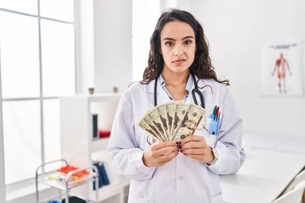 Young doctor woman holding money clueless and confused expression. doubt concept.