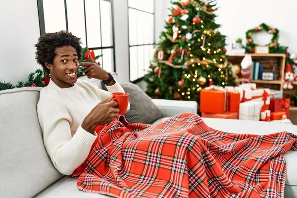 年轻的非洲裔美国人坐在沙发上 靠着圣诞树喝咖啡 手指点点着脸和鼻子 笑容满面 美的概念 — 图库照片