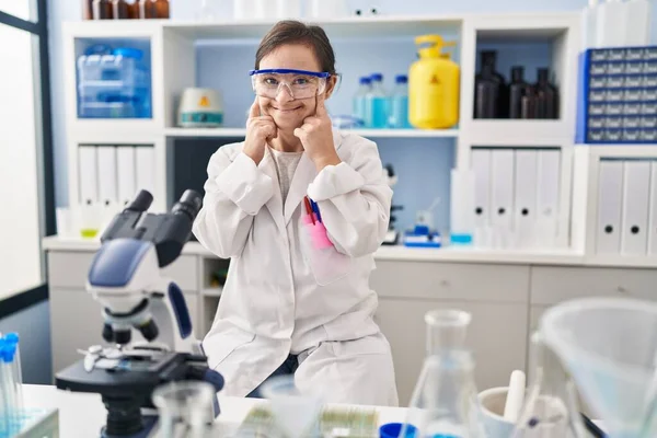 Hispanic girl with down syndrome working at scientist laboratory smiling with open mouth, fingers pointing and forcing cheerful smile