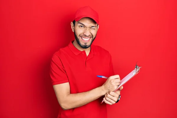 Hispanic Man Beard Wearing Courier Uniform Holding Clipboard Winking Looking — Stockfoto