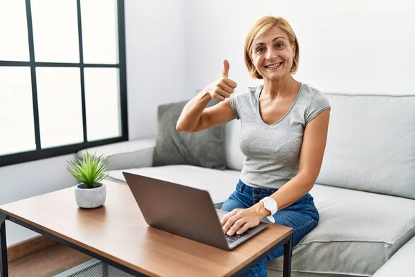 Middle age blonde woman using laptop at home doing happy thumbs up gesture with hand. approving expression looking at the camera showing success.