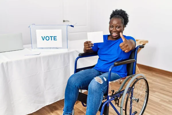 Young african woman sitting on wheelchair voting putting envelop in ballot box approving doing positive gesture with hand, thumbs up smiling and happy for success. winner gesture.