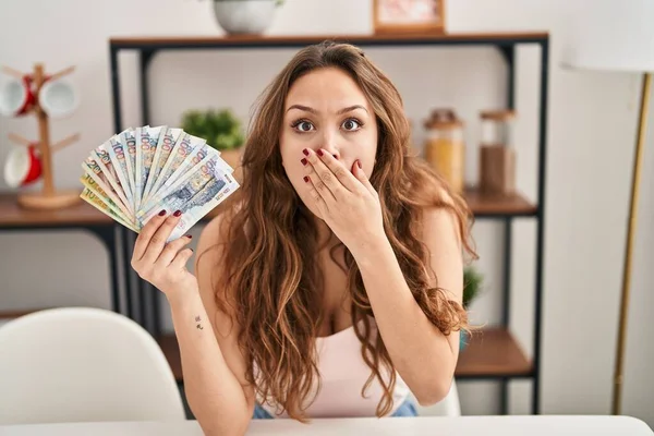 Young hispanic woman holding peruvian sol banknotes covering mouth with hand, shocked and afraid for mistake. surprised expression