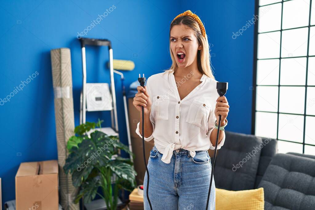 Young blonde woman moving to a new home holding plugs angry and mad screaming frustrated and furious, shouting with anger. rage and aggressive concept. 
