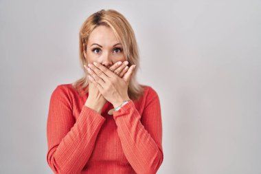 Blonde woman standing over isolated background shocked covering mouth with hands for mistake. secret concept. 