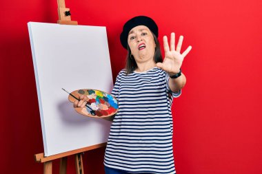 Middle age hispanic woman standing drawing with palette by painter easel stand doing stop gesture with hands palms, angry and frustration expression 