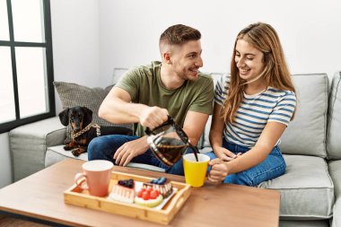 Genç İspanyol çift mutlu bir şekilde gülümsüyor. Evde köpekle birlikte kanepede oturuyorlar..