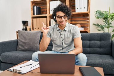 Kıvırcık saçlı İspanyol adam danışma ofisinde online seans yapıyor. Elini ve parmağını yana uzatarak mutlu bir şekilde gülümsüyor. 
