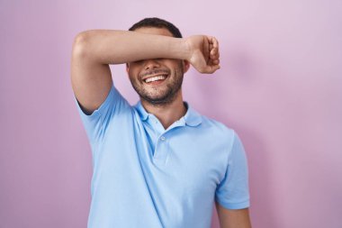 Hispanic man standing over pink background covering eyes with arm smiling cheerful and funny. blind concept. 