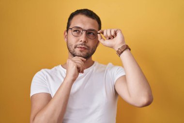 Hispanic man holding glasses serious face thinking about question with hand on chin, thoughtful about confusing idea 