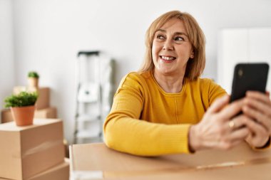 Orta yaşlı sarışın kadın yeni evinde akıllı telefon kullanarak mutlu bir şekilde gülümsüyor..