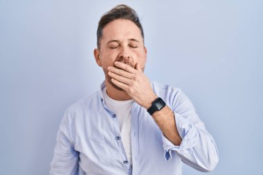 Middle age caucasian man standing over blue background bored yawning tired covering mouth with hand. restless and sleepiness. 