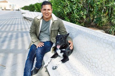 Genç Latin adam gülümsüyor. Parkta köpekle bankta mutlu mesut oturuyor..