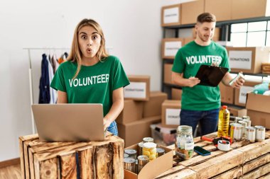 Young woman and man wearing volunteer t shirt at donations stand scared and amazed with open mouth for surprise, disbelief face 