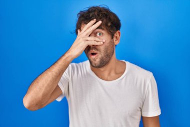 Hispanic young man standing over blue background peeking in shock covering face and eyes with hand, looking through fingers with embarrassed expression. 