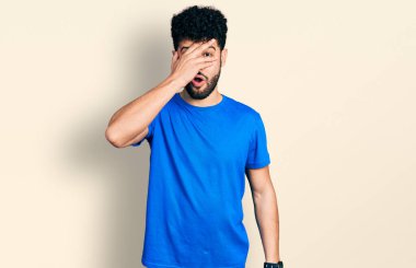 Sakallı genç, mavi tişörtlü bir Arap adam. Şok olmuş bir şekilde yüzünü ve gözlerini kapatıyor. Utanmış bir ifadeyle parmaklarından bakıyor.. 