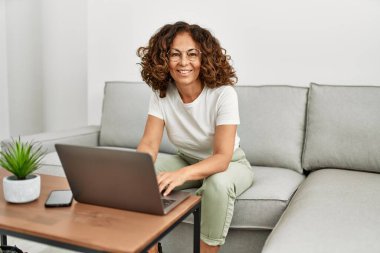 Orta yaşlı İspanyol kadın evde dizüstü bilgisayar kullanarak gülümsüyor.