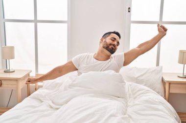 Young hispanic man stretching arms sitting on bed at bedroom