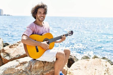 Genç İspanyol adam klasik gitar çalıyor plajda rock 'ın üzerinde oturuyor..