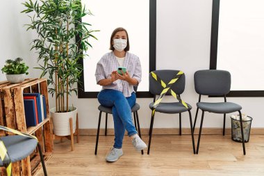 Middle age hispanic woman wearing safety mask using smartphone at waiting room