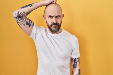 Young hispanic man with tattoos standing over yellow background confuse and wondering about question. uncertain with doubt, thinking with hand on head. pensive concept. 