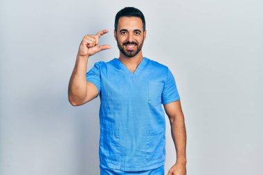 Sakallı yakışıklı, mavi erkek hemşire üniforması giyen, gülümseyen, kendine güvenen, el hareketi yapan, parmaklarıyla işaret yapan ve kameraya bakan bir İspanyol. ölçüm kavramı. 