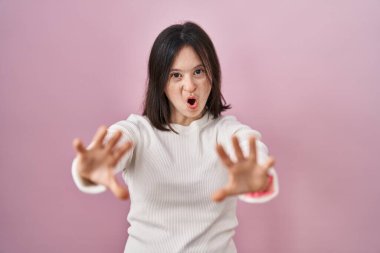 Woman with down syndrome standing over pink background doing stop gesture with hands palms, angry and frustration expression 