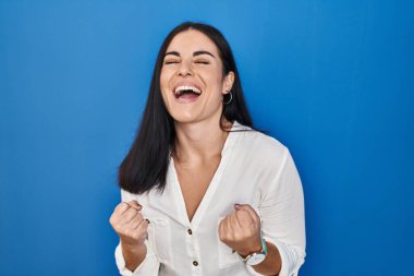 Young hispanic woman standing over blue background very happy and excited doing winner gesture with arms raised, smiling and screaming for success. celebration concept. 