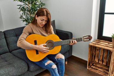 Orta yaşlı beyaz kadın klasik gitar çalıyor. Evdeki kanepede oturuyor..
