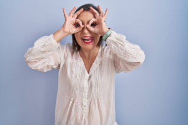 Middle age hispanic woman standing over blue background doing ok gesture like binoculars sticking tongue out, eyes looking through fingers. crazy expression. 