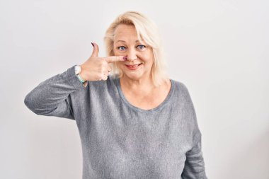 Middle age caucasian woman standing over white background pointing with hand finger to face and nose, smiling cheerful. beauty concept 
