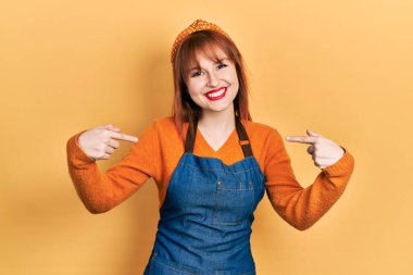 Önlük giyen, kendine güvenen, gülümseyen, kendini gururlu ve mutlu gösteren kızıl saçlı genç bir kadın.. 