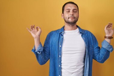 Hispanic man standing over yellow background relaxed and smiling with eyes closed doing meditation gesture with fingers. yoga concept. 