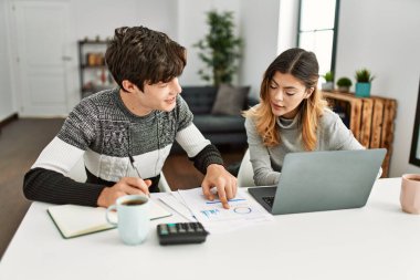 Genç çift evde masada dizüstü bilgisayarla çalışırken mutlu bir şekilde gülümsüyor..