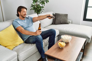 Orta yaşlı beyaz bir adam evde televizyon izlerken kendinden emin gülümsüyor.