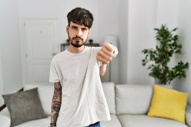 Hispanic man with beard at the living room at home looking unhappy and angry showing rejection and negative with thumbs down gesture. bad expression. 