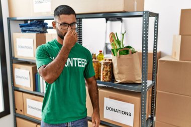 Young indian man volunteer holding donations box smelling something stinky and disgusting, intolerable smell, holding breath with fingers on nose. bad smell 