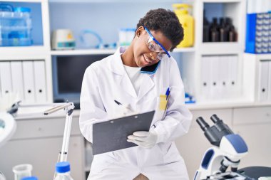 Bilim adamı üniforması giyen Afrikalı Amerikalı kadın. Akıllı telefondan konuşuyor. Laboratuvarda panoya yazı yazıyor.